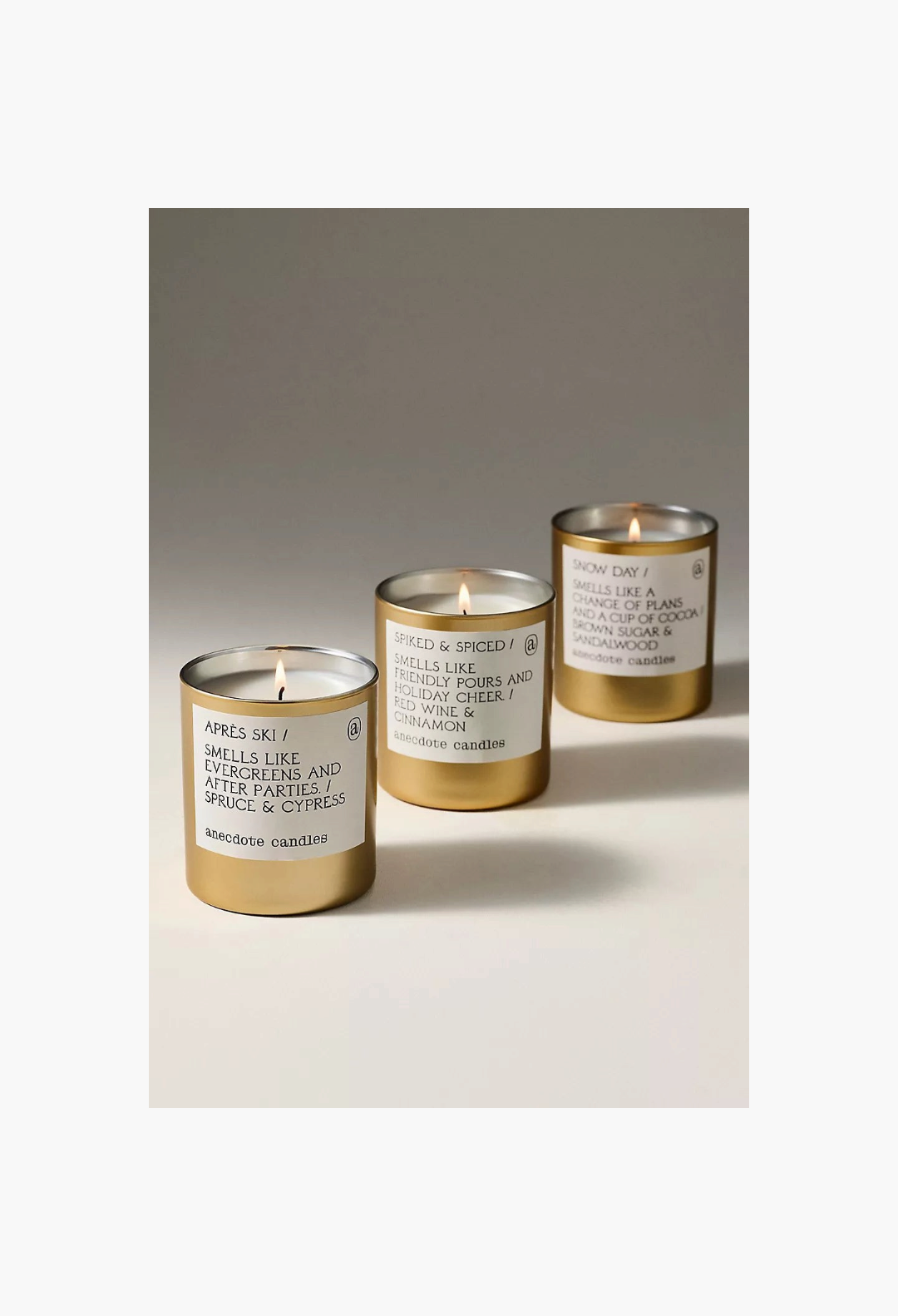 GLAM MODA Anecdote Gourmand candles in gold glass vessels with white labels and lit wicks on a neutral surface.