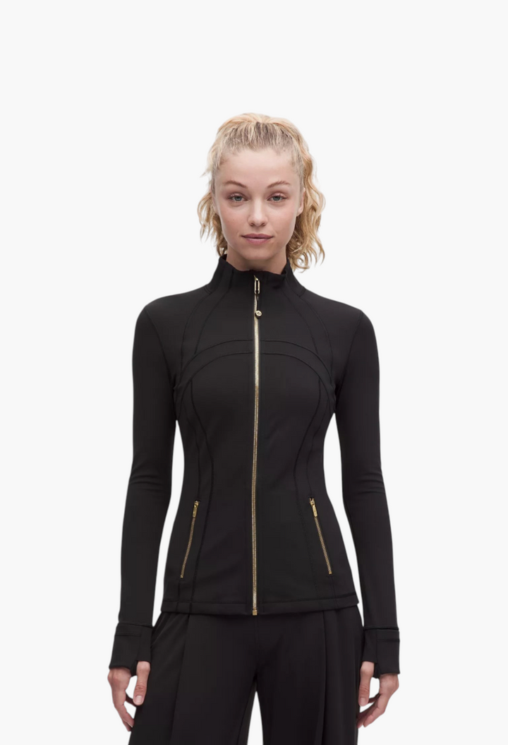GLAM MODA Define Jacket Nulu in black with gold zippers, thumbholes, and a slim-fit athletic design on a model.