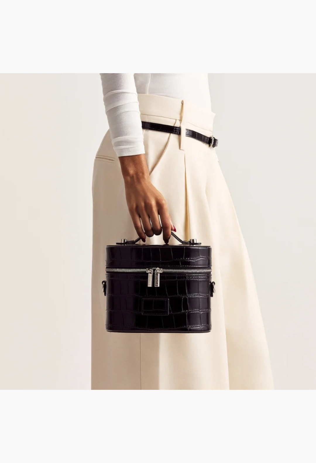 GLAM MODA black vegan leather oval toiletry case with crocodile texture held by a model in cream trousers.
