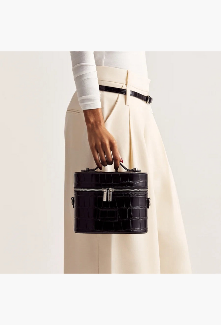 GLAM MODA black vegan leather oval toiletry case with crocodile texture held by a model in cream trousers.