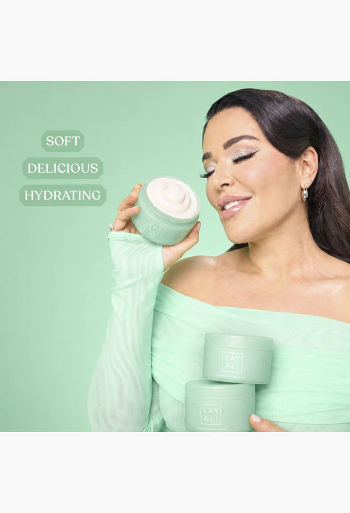 GLAM MODA model holding KAYALI Yum Pistachio Silk Soufflé body cream jars against a soft green background.