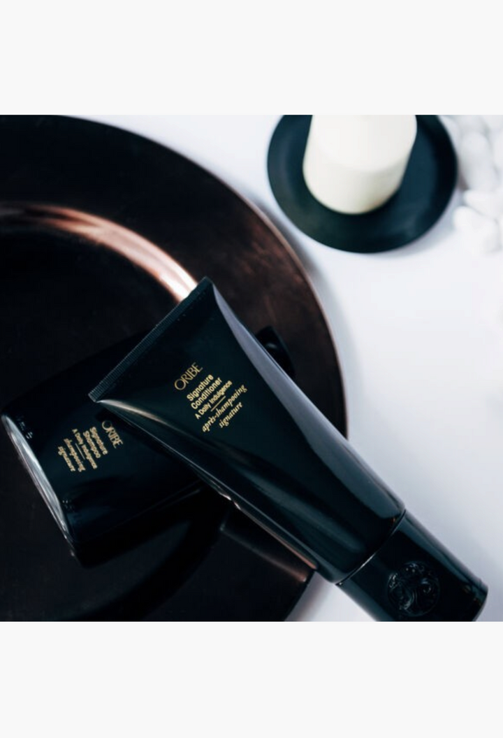 GLAM MODA Oribe Signature Shampoo and Conditioner in black tubes on a dark reflective surface.