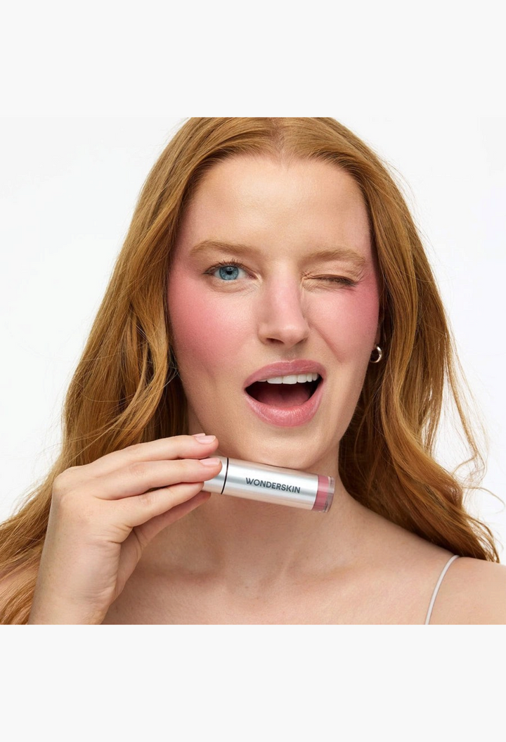 GLAM MODA model winking while holding Wonderskin Wonder Blading All-Day Blush Stain with a natural pink cheek flush
