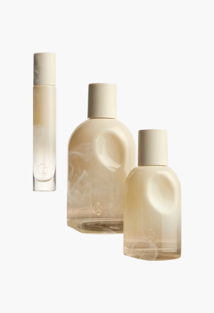 GLAM MODA Glossier You Doux woody eau de parfum bottles in various sizes displayed on a textured wooden surface.