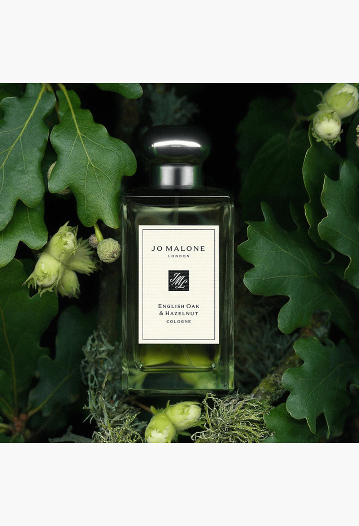 GLAM MODA Jo Malone London English Oak & Hazelnut cologne bottle nestled among fresh green oak leaves and hazelnuts.