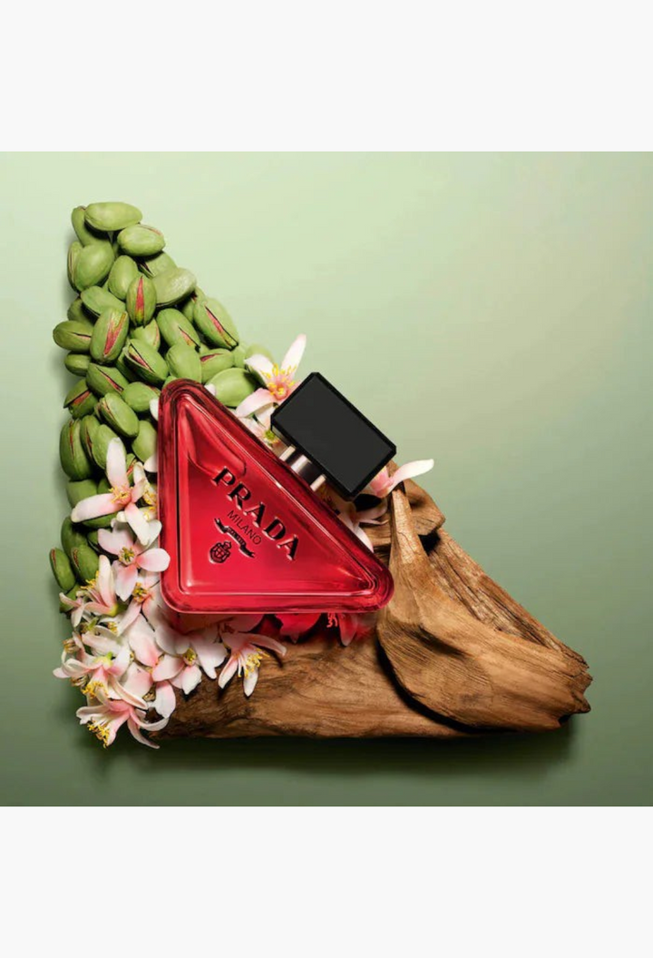GLAM MODA Prada Paradoxe perfume bottle in a triangular red glass design, surrounded by green pistachios and flowers.
