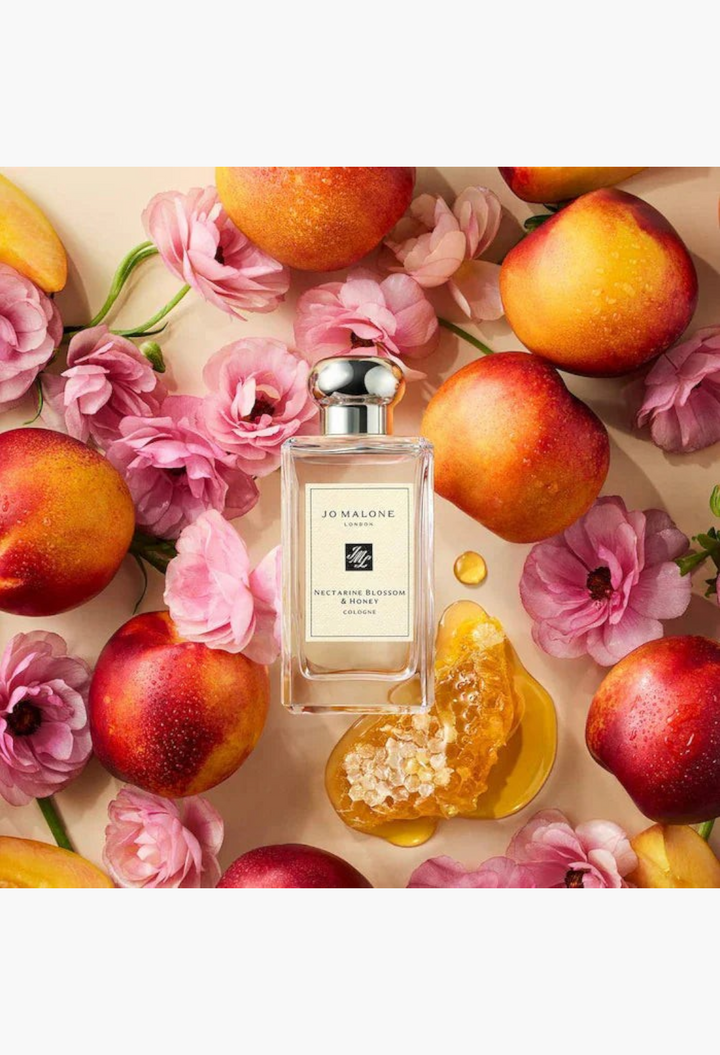 GLAM MODA Jo Malone London Nectarine Blossom & Honey cologne bottle surrounded by fresh nectarines and pink flowers.
