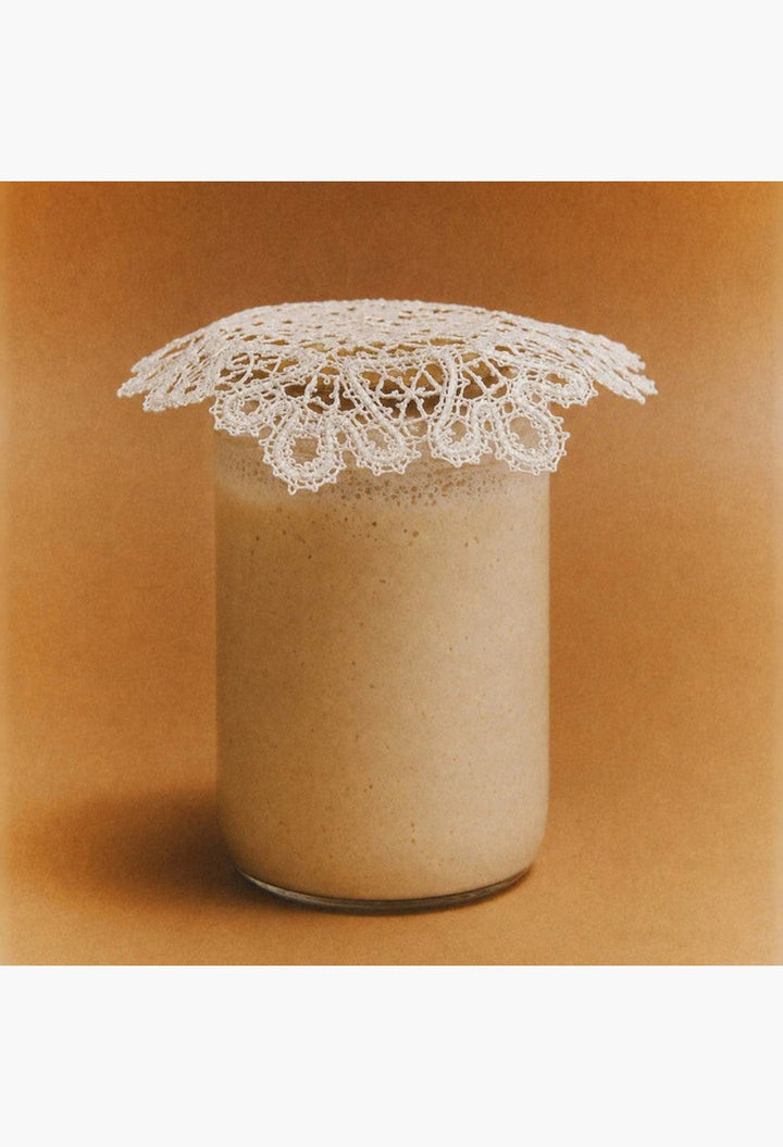 GLAM MODA Le Chouchou peptide lip balm in a clear jar topped with a delicate white lace doily on a tan background.