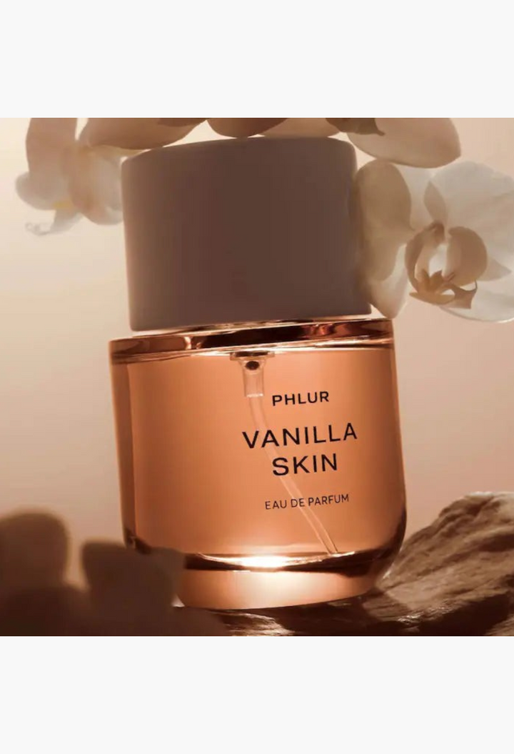 GLAM MODA Phlur Vanilla Skin Eau de Parfum bottle with warm amber liquid, white orchid flowers, and stone base.
